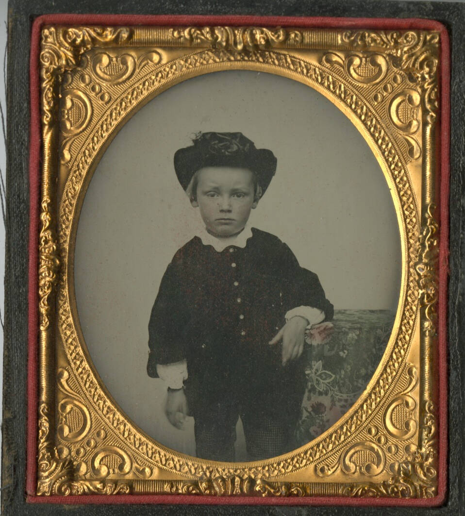 Black and white ambrotype photographic portrait of a young child in a dark outfit with a hat.